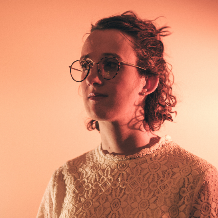 An image of a woman with curly hair and round glasses. She wears a lace top and is looking away from the camera.
